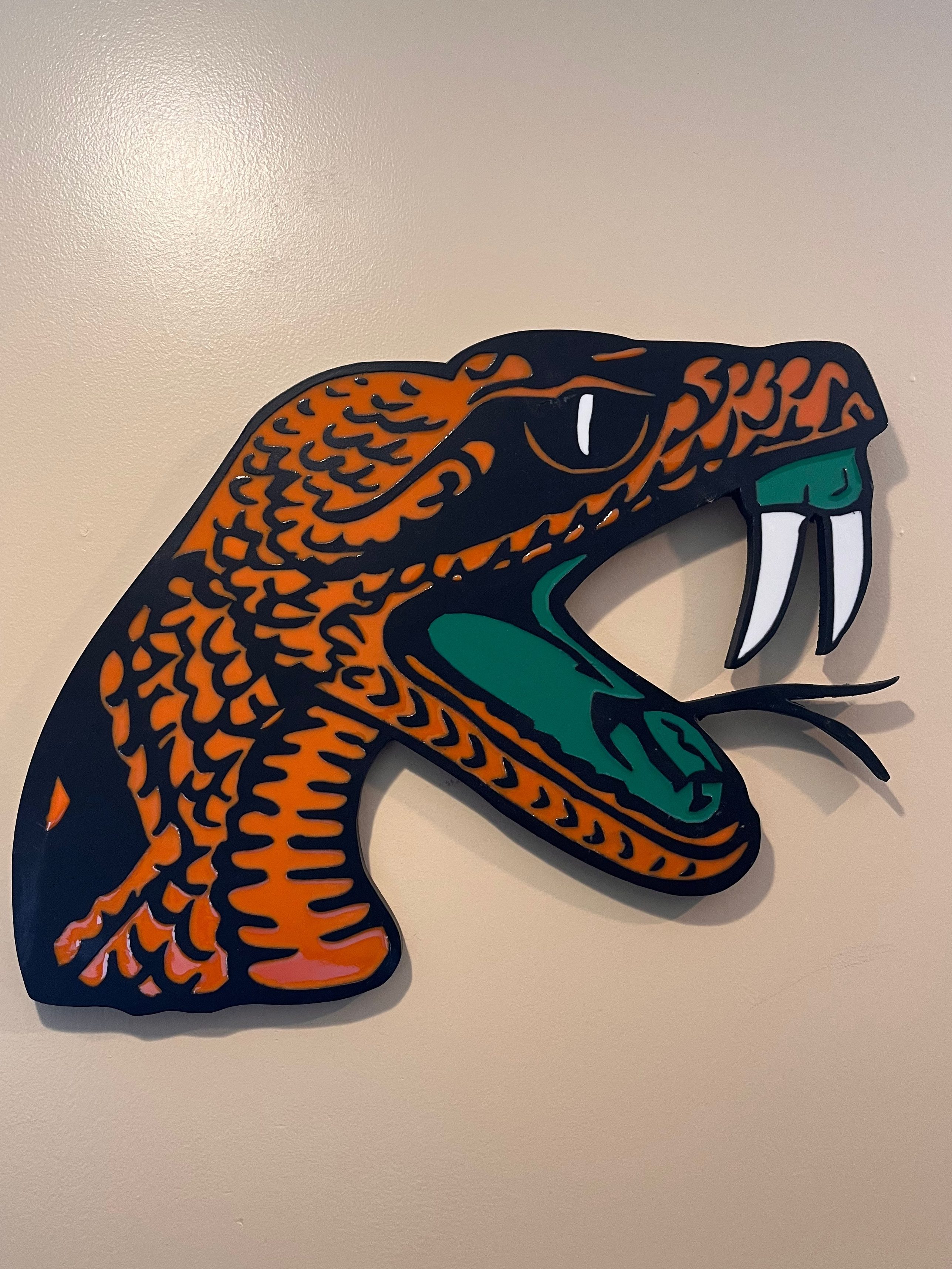 Colorful wall art depicting the head of a snake with striking features, including fangs and an open mouth, set against a neutral wall background.